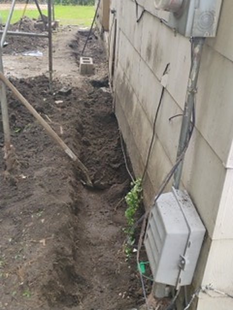 A trench dug along the outside of a house foundation, with the dirt piled on the lawn